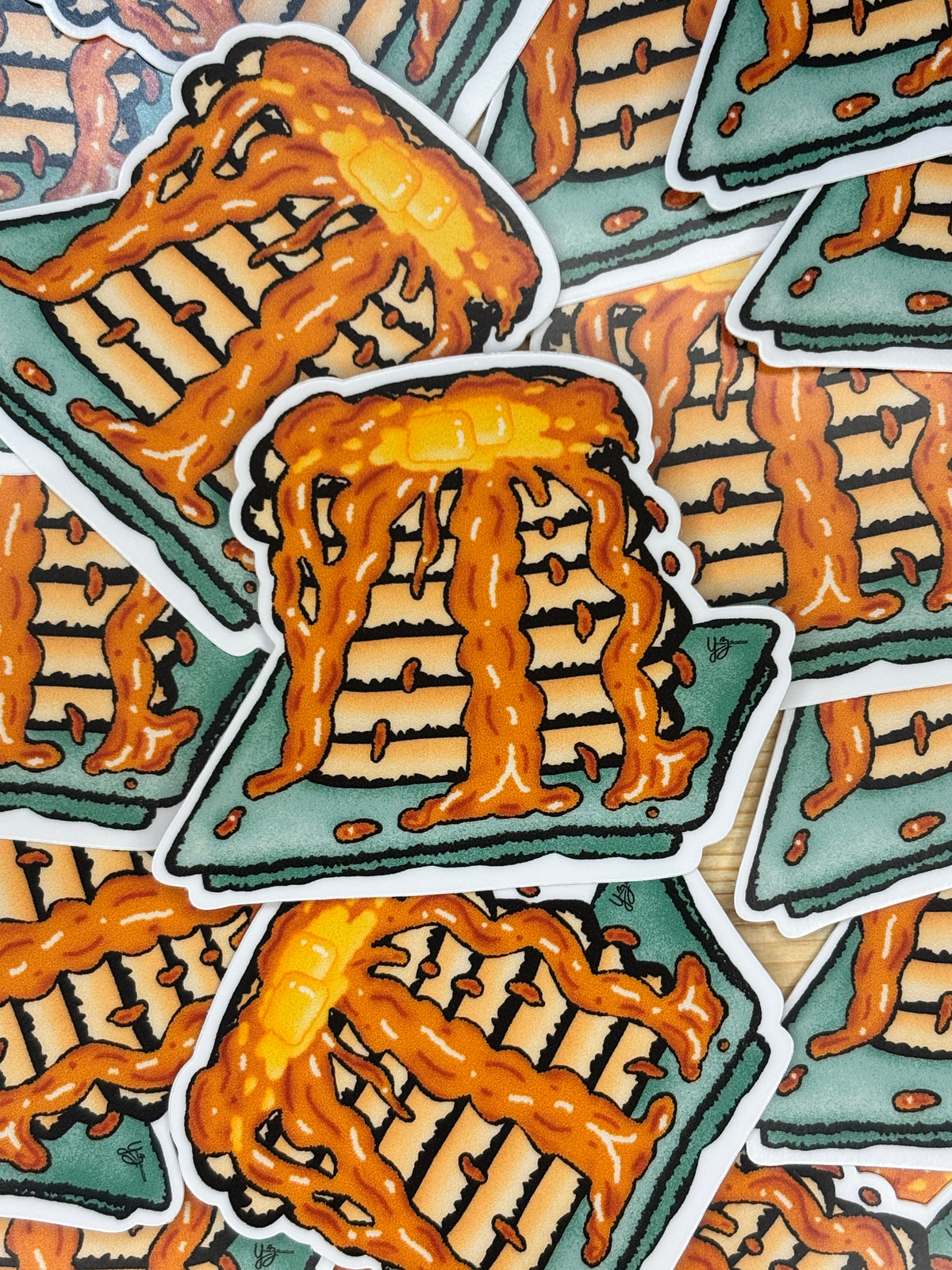 A close-up of multiple illustrated food stickers arranged on a light wood surface. The stickers depict rectangular pastries or cakes with tan layers, dark brown crumb lines, and orange-brown drizzle on a green base.