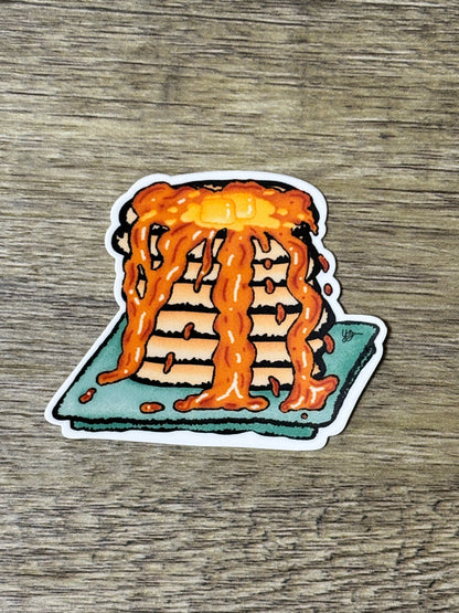 A die-cut sticker featuring a hand-drawn stack of pancakes dripping with maple syrup, placed on a teal plate against a wood-grain background.
